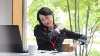 オフィスで肩をさする女性。冷えや姿勢の偏りによる肩こりの例